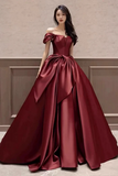 Woman wearing a burgundy off-shoulder gown with a large bow detail against a neutral wall.