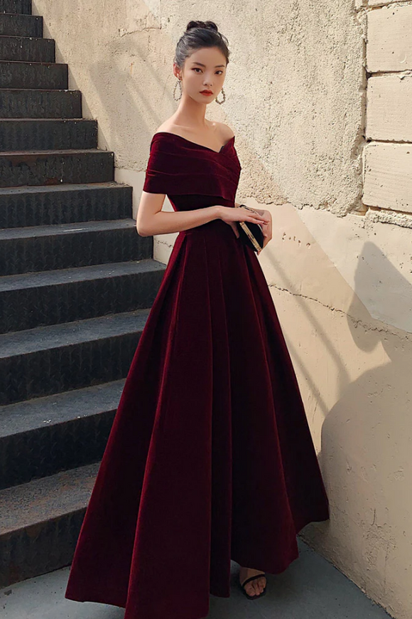 Woman in a burgundy off-shoulder dress standing on steps against a textured wall.