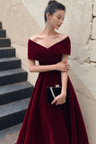 Woman in a burgundy off-shoulder dress holding a clutch against a textured wall.
