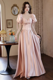 Woman in a long, light pink dress standing in an elegant room with a table and decorative items.