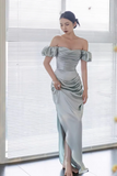 Woman wearing a silver off-shoulder gown standing in front of a window with blinds.