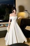 Woman in a white off-shoulder dress standing indoors with a cityscape view.