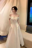 Woman in a white off-shoulder dress standing in a room with a couch and window.
