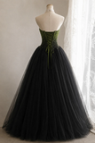 A strapless ball gown displayed on a mannequin, featuring a structured olive green corset-style bodice with beadwork and lace-up detailing, paired with a voluminous black tulle skirt, set against a neutral indoor background with a window curtain.