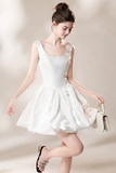 A woman modeling a short white satin dress with a square neckline and a flared skirt, holding a small white handbag against a soft beige studio background.
