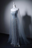 A floor-length formal gown displayed on a mannequin in a dim studio setting. The dress has a structured corset-style bodice with dense bead and sequin embellishment, a sheer draped cape overlay, and a long flowing train.