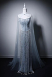 A floor-length formal gown displayed on a mannequin against a dark gray studio background. The dress has a fitted embellished bodice, a sheer draped cape-like overlay, and a long flowing train in pale icy blue and silver tones.