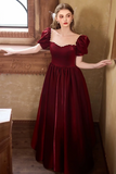 A woman wearing a long burgundy velvet gown with puff sleeves, a square neckline trimmed with gold lace, and a full skirt, posing in a rustic indoor setting with textured walls and a window.