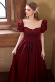 A woman wearing a floor-length burgundy gown with puff sleeves, a square neckline trimmed with gold lace, and a full skirt, standing in a warm-toned indoor setting.