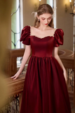 A woman wearing a deep burgundy red gown with puffed sleeves, a sweetheart neckline trimmed with gold lace, and a full skirt, standing in an indoor setting with warm lighting.