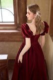 A woman wearing a deep burgundy velvet gown with puff sleeves, a sweetheart neckline, and lace-up corset detailing, standing in a warm-toned indoor setting.