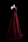 A floor-length burgundy velvet gown displayed on a white mannequin against a solid black background. The strapless gown features a ruched bodice with a gold embellished waistband and a full, flowing skirt.
