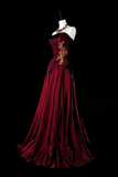 A floor-length burgundy evening gown displayed on a white mannequin against a solid black background. The gown features a strapless neckline, a ruched bodice with gold floral embroidery, and a full, flowing skirt.