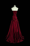 A strapless floor-length gown in deep burgundy red, displayed on a white mannequin torso against a solid black background. The dress features a ruched bodice and a full, flowing skirt.