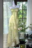 A pale yellow satin evening gown with green floral embroidery and a sheer draped tie at the waist, displayed on a white mannequin torso in a sunlit room near a window.
