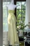 A pale yellow strapless maxi dress displayed on a white mannequin in front of a window with outdoor greenery visible, with a side table and flowers nearby.