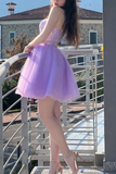 A woman wearing a short lavender tulle prom dress with a sparkling embellished bodice, posing on an outdoor balcony with a stone building in the background.