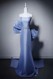 A light blue satin evening gown displayed on a white mannequin against a dark gray studio background. The dress features a strapless neckline, dramatic oversized bow details on the shoulders, a jeweled embellished waist, and a long train with a side panel.