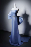 A floor-length light blue evening gown displayed on a white mannequin in a studio setting. The dress has a fitted bodice, a dramatic draped one-shoulder design with a large flowing sleeve, a jeweled embellishment at the waist, and a long train.
