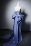 A blue satin gown displayed on a white mannequin against a dark gray studio wall, featuring a dramatic oversized bow at the back with a long train.