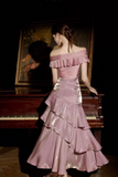 A woman stands with her back to the camera wearing an off-the-shoulder, tiered pink gown with a metallic sheen, posed in front of a dark room and a large framed painting near a piano.