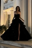 A woman wearing a strapless black ball gown with a high thigh slit and sparkling embellishments, posing on the steps of a building with warm white columns.