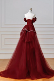 A floor-length off-the-shoulder ball gown in deep burgundy red tulle, displayed on a white mannequin torso against a white paneled wall and light wood floor.