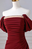 A close-up of a burgundy off-the-shoulder evening gown displayed on a white mannequin torso. The dress features a sweetheart neckline embellished with a line of clear rhinestones over a beige underlayer, with ruched draping across the bodice and skirt.