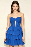 A woman modeling a strapless royal blue mini dress with a sweetheart neckline, twisted bodice detail, and a tiered ruffle skirt against a plain cream background.