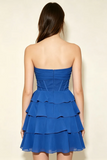 A woman viewed from the back wearing a strapless royal blue mini dress with a ruched bodice and tiered ruffle skirt, against a plain light beige studio background.