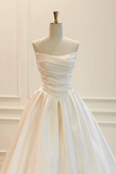 A strapless white satin ball gown with a pleated bodice and full skirt, displayed on a cream mannequin against a light cream paneled wall.