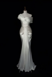 An off-the-shoulder floor-length white gown with draped satin-like fabric and a gathered waist detail, displayed on a mannequin against a black background.