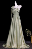 A floor-length formal gown displayed on a white mannequin against a black background. The dress has an asymmetrical draped neckline with a bow detail, a fitted waist, and a full satin-like skirt with a slight train.