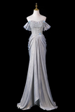 A floor-length off-the-shoulder evening gown in a shimmering light silver-gray fabric, featuring a heavily embellished beaded bodice with small blue accents and a draped skirt, displayed on a mannequin against a solid black background.