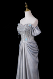 A formal off-the-shoulder gown displayed on a mannequin against a black background. The dress is a shimmering light silver-gray with a heavily embellished bodice covered in rhinestones and crystals, featuring light blue bead accents and a draped skirt.