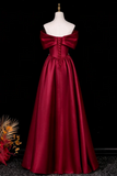 A floor-length off-the-shoulder red satin gown displayed on a white mannequin against a black background, with a large bow at the neckline and lace-up front detailing.