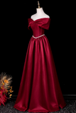 A floor-length off-the-shoulder formal gown in deep burgundy satin, displayed on a white mannequin against a black background. The dress features a large bow detail at the neckline, a fitted bodice, and a thin silver rhinestone waistband.