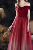 A woman wearing a floor-length off-the-shoulder evening gown in deep red, with a structured bodice, sheer tulle overlay, and embellished front detail, posed against a dark textured backdrop with dried grass decor.