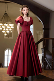 A woman stands in an elegant indoor setting wearing a floor-length burgundy gown with puff sleeves, a square neckline, and a full skirt.
