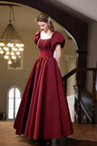 A woman wearing a floor-length burgundy ball gown with puffed short sleeves, a square neckline, and a full skirt, standing in an indoor staircase setting with a chandelier visible in the background.