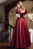 A woman wearing a floor-length burgundy ball gown with puff sleeves and a lace-up back, standing on an indoor staircase in a dimly lit elegant room.