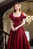 A woman wearing a floor-length burgundy gown with puffed sleeves and a square neckline, standing in an elegant indoor setting with a staircase and chandelier in the background.